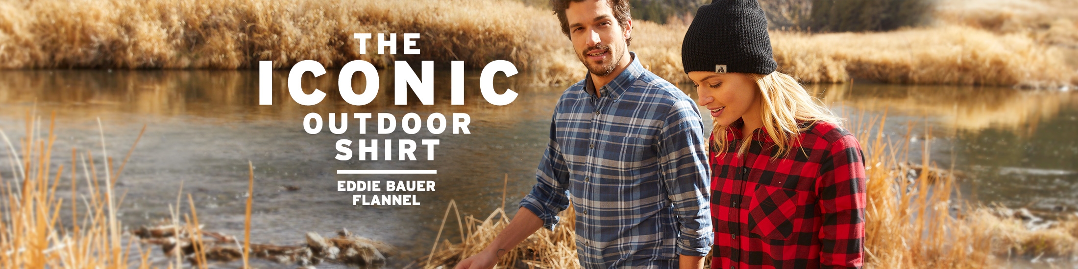 Two people wearing flannel shirts walk through fall foliage next to a river